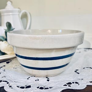 May include: A cream-colored ceramic mixing bowl featuring two horizontal navy blue stripes encircling the exterior. The bowl has a wide, thick rim and a rounded base, showcasing a smooth, glazed finish throughout its entire surface area.