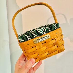 May include: A small, woven basket with a wooden handle and a green and white striped fabric lining. The basket has a rectangular shape and a small metal tag that reads "LONGABERGER 1998 Hostess Appreciation Basket."