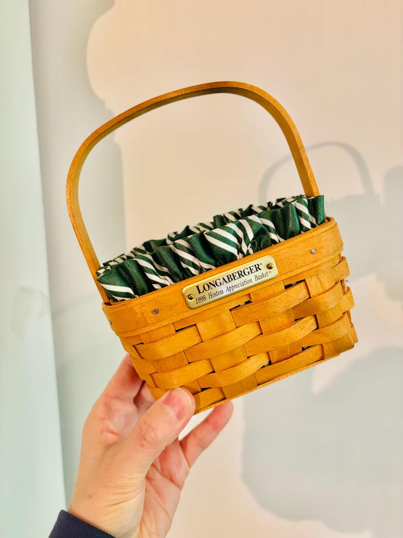 May include: A small, woven basket with a wooden handle and a green and white striped fabric lining. The basket has a rectangular shape and a small metal tag that reads "LONGABERGER 1998 Hostess Appreciation Basket."