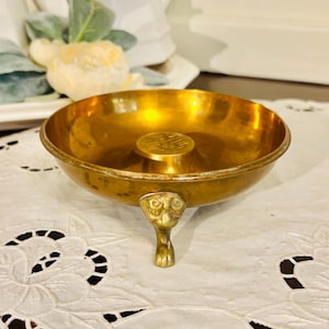 May include: A small, antique brass bowl with a single ornate foot. The bowl has a circular design in the center and a shiny, golden finish. It sits on a white lace tablecloth, suggesting a decorative or display item.