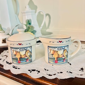 May include: Cream-colored ceramic creamer and sugar bowl set. Each piece features a blue-bordered rectangular design with a cartoon cow on a rocking horse and the word "COW". The set is on a white lace doily.
