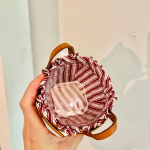 May include: A small, round basket with a clear glass insert. The basket has a red and white striped fabric lining and a brown leather handle. The basket is held in a person's hand.