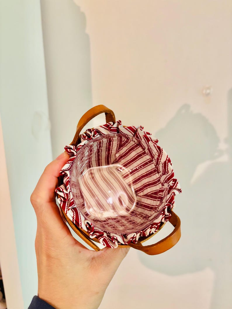 May include: A small, round basket with a clear glass insert. The basket has a red and white striped fabric lining and a brown leather handle. The basket is held in a person's hand.