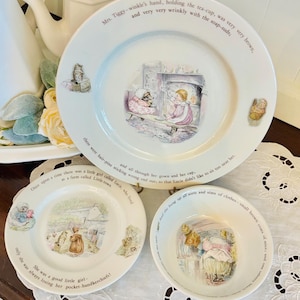 May include: A set of three white ceramic plates and a bowl with illustrations and text from a children's story. The plates and bowl feature images of characters and scenes, with text around the edges.