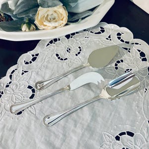 May include: Three silver serving utensils: a cake server, a cheese knife, and a serving spoon, arranged on a white, floral-embroidered doily. The utensils have beaded edges and are displayed near a white serving dish with a floral arrangement.