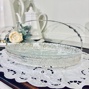 May include: A silver-toned serving dish with a clear glass insert and a decorative handle. The dish sits on a white, embroidered doily. Teapots and a faux flower arrangement are in the background.