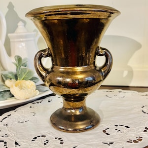 May include: A bronze-colored vase with a wide, fluted top and two curved handles. The vase has a pedestal base and sits on a white, lace-trimmed doily. The vase is likely made of ceramic or metal and has a glossy finish.