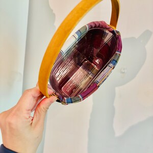 May include: A small, plaid-lined basket with a curved, yellow handle. The basket contains a clear container with several items inside. The plaid fabric features red, blue, and purple hues. The handle is made of a smooth, light-colored material.
