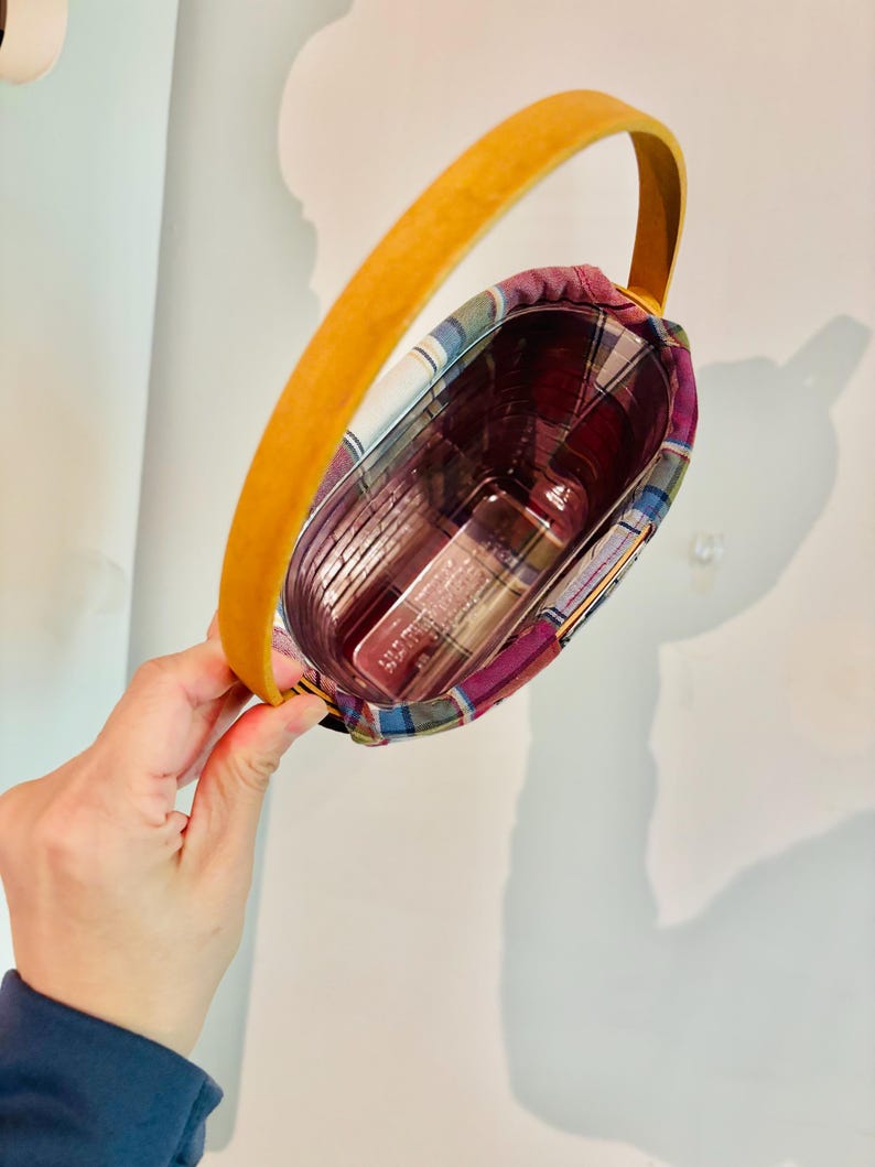May include: A small, plaid-lined basket with a curved, yellow handle. The basket contains a clear container with several items inside. The plaid fabric features red, blue, and purple hues. The handle is made of a smooth, light-colored material.