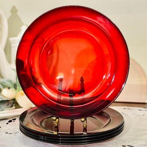May include: A stack of red glass plates, with a matching plate on top, displayed on a decorative stand. The plates have a glossy finish and are arranged on a white lace tablecloth. The plates are a vibrant red color.