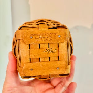 May include: A small, square, handwoven basket made of light brown wood. The basket has a woven design and is held in a hand. The text "LONCABERGER BASKETS HANDWOVEN DRESDEN OHIO USA" is visible. The basket is a vintage item.
