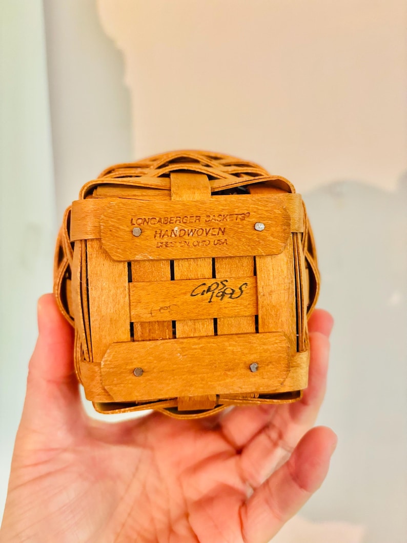 May include: A small, square, handwoven basket made of light brown wood. The basket has a woven design and is held in a hand. The text "LONCABERGER BASKETS HANDWOVEN DRESDEN OHIO USA" is visible. The basket is a vintage item.
