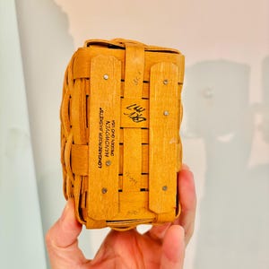May include: A small, rectangular Longaberger basket, handwoven from light brown wood. The basket has a lid and is held together with wooden slats and metal rivets. The text "LONGABERGER BASKETS HANDWOVEN DRESDEN, OHIO USA" is visible.