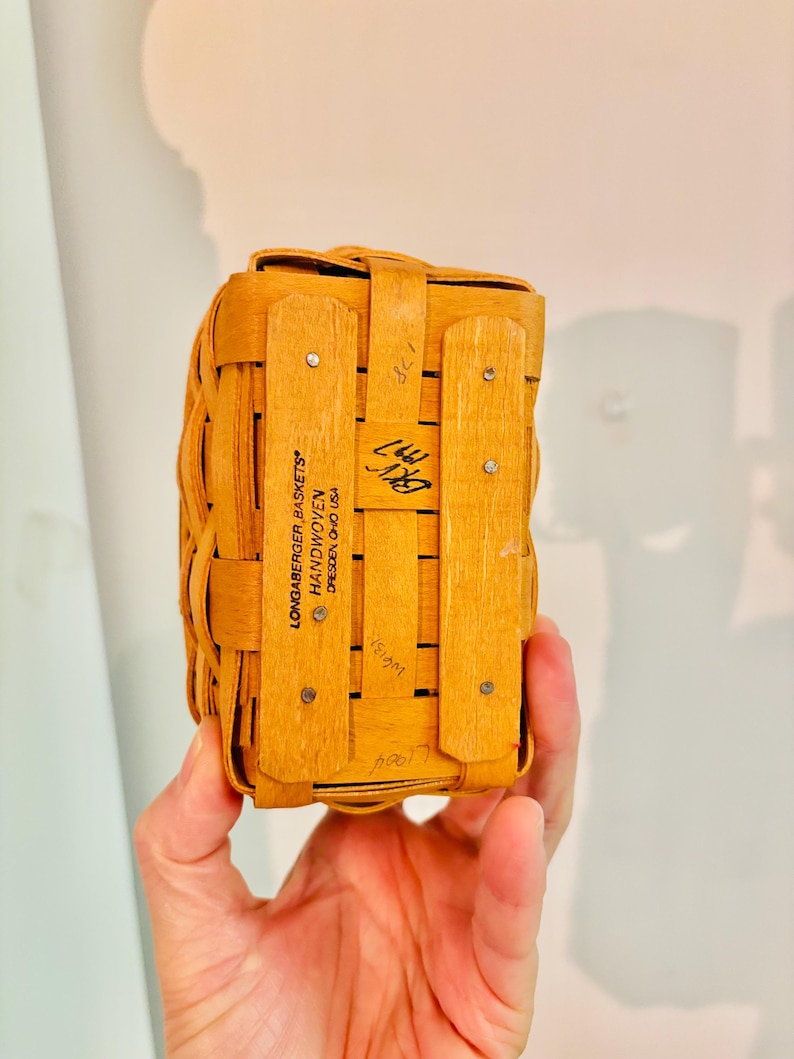 May include: A small, rectangular Longaberger basket, handwoven from light brown wood. The basket has a lid and is held together with wooden slats and metal rivets. The text "LONGABERGER BASKETS HANDWOVEN DRESDEN, OHIO USA" is visible.