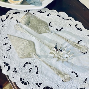 May include: Silver serving utensils with ornate floral designs are arranged on a white, lace-edged doily. The set includes a cake server, a pie server, and a spaghetti server. The background features a white serving dish and floral arrangement.