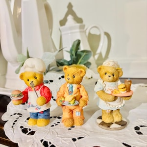 May include: Three charming bear figurines in chef and server attire. One bear holds a burger, another a plate of food, and the third a tray with a burger and drink. The bears are on a white lace tablecloth, with white teapots in the background.