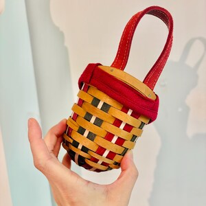 May include: A small, woven basket with a red handle and rim. The basket features a pattern of red, black, and natural wood-toned strips. The handle is a deep red, and the basket is held in a person's hand.