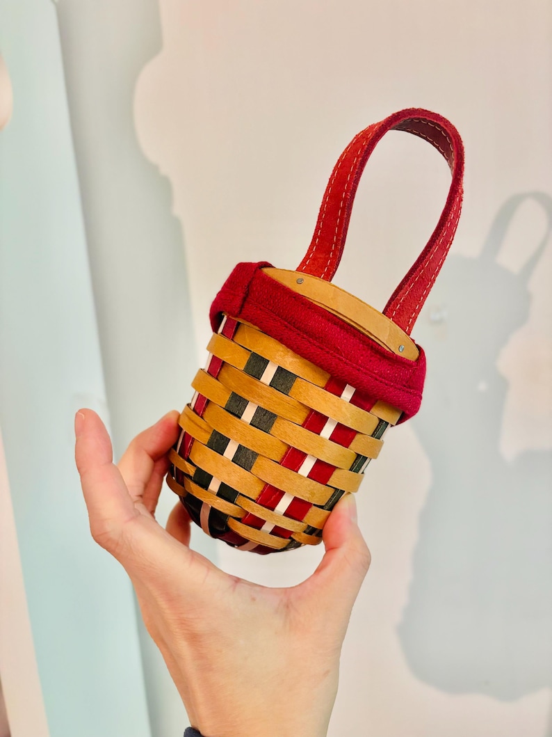 May include: A small, woven basket with a red handle and rim. The basket features a pattern of red, black, and natural wood-toned strips. The handle is a deep red, and the basket is held in a person's hand.