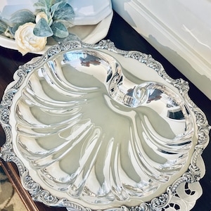 May include: A silver-plated serving dish with a shell-like design. The dish has a scalloped edge and a handle. It is sitting on a white doily on a dark wooden surface. A white teapot is in the background.