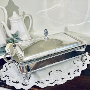 May include: A rectangular silver-plated serving dish with a polished finish, featuring two side handles, four ornate feet, and a removable lid topped with a decorative finial shaped like a small pineapple. The dish has a sleek, reflective surface.