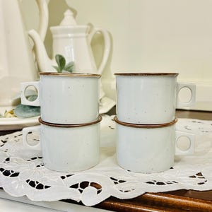 May include: Four white ceramic mugs with a speckled finish and brown rims are stacked on a white lace doily. The mugs have square handles. A white teapot and other teacups are in the background.