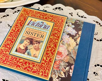 May include: A vintage book titled "Victoria No Friend Like A Sister" with a red and gold cover. The book is open, revealing a floral patterned interior and a blue spine. The book is resting on a white lace doily.