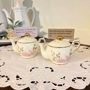 May include: Two small, cream-colored teapots with gold accents and floral designs. Each teapot has a gold knob on the lid and a curved handle. The text on the cards reads: "Courage is knowing what not to fear." and "Everything has beauty, but not everyone sees it."