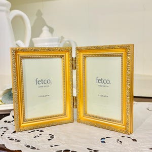 May include: A gold-colored, hinged picture frame with ornate detailing. Each frame displays the text "fetco. HOME DECOR" and the dimensions "3 1/2 in. x 5 in.". The frame is set on a white lace doily.