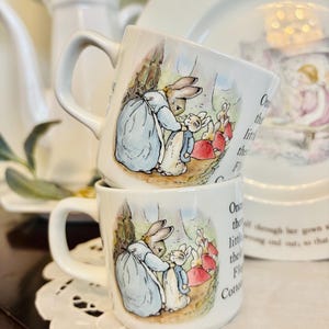 May include: Two white ceramic mugs stacked on top of each other, each featuring a colorful illustration of rabbits in a forest setting. The mugs have curved handles and are decorated with text from a children's story.