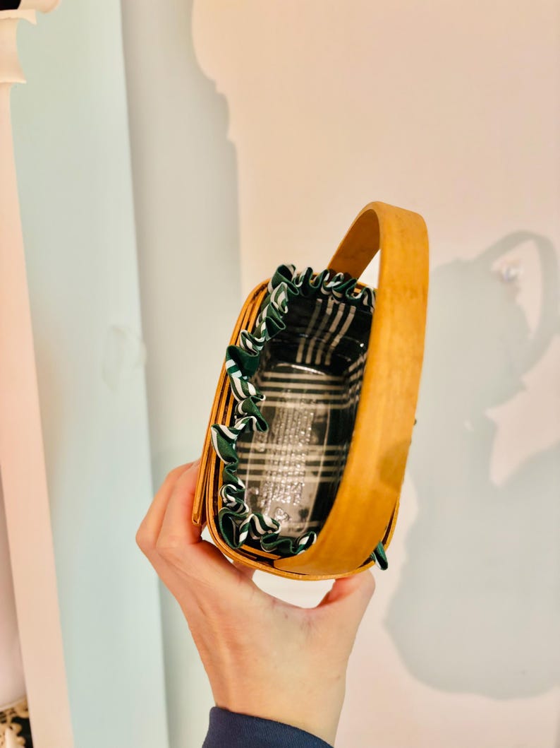May include: A small, light brown wicker basket with a curved wooden handle. The interior features a black and white plaid fabric lining, trimmed with green and white ribbon. The basket is designed for carrying small items.