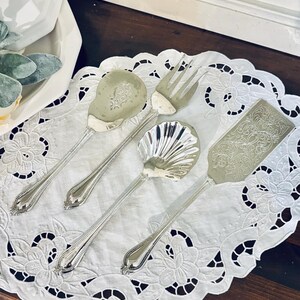 May include: A set of four silver-toned serving utensils including a solid spoon, a three-tined fork, a shell-shaped spoon, and a rectangular server. Each piece features a long, slender handle with a beaded edge and a decorative floral pattern.
