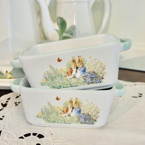 May include: Two rectangular white ceramic dishes with light blue handles and rims. Each dish features a colorful illustration of two rabbits in a garden setting with butterflies. The dishes are stacked on a white lace tablecloth.