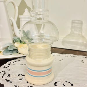 May include: A ceramic oil lamp base with a cream-colored finish, featuring two horizontal blue stripes and one pink stripe around the center. The lamp includes a clear, rounded glass chimney top that sits securely on the cylindrical ceramic body.