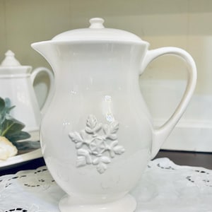 May include: A white ceramic pitcher with a lid and a handle, featuring a raised, embossed snowflake design on the front. The pitcher has a curved body, a flared spout, and a pedestal base, all finished in a smooth, glossy white glaze.