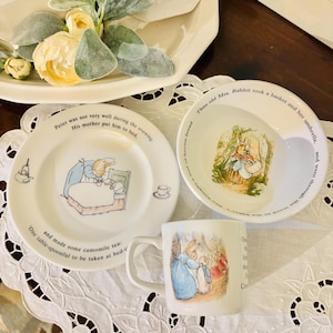 May include: A white ceramic dish, bowl, and mug set featuring illustrations of Peter Rabbit. The plate has text that reads, "Peter was not very well during the evening. His mother put him to bed." The bowl and mug also have text.