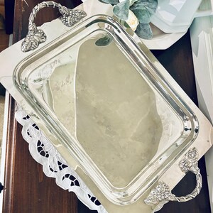 May include: A rectangular silver serving tray with ornate handles. The tray is placed on a white lace doily and a wooden surface. Decorative floral elements and white teacups are visible in the background.