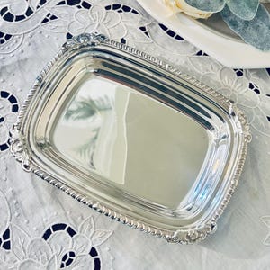 May include: A rectangular silver-toned tray with a polished, reflective surface and a decorative, raised border featuring intricate scrollwork and beaded detailing along the edges. The tray has rounded corners and a smooth, flat center area.