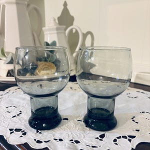 May include: Two clear glass goblets with a dark teal base and stem sit on a white lace doily. The goblets have a rounded bowl shape. The background includes white ceramic teapots and a white wall.