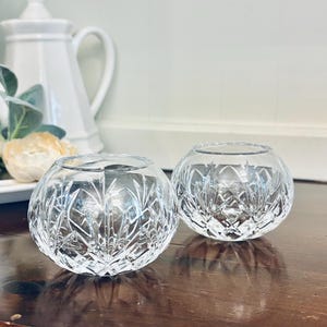 May include: Two clear, round glass votive candle holders with intricate cut crystal designs. The candle holders sit on a dark wooden surface, with a white teapot and decorative items in the background.