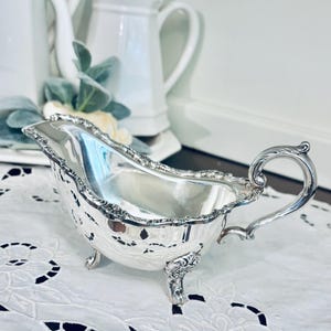 May include: A silver-plated gravy boat featuring an ornate, pierced lattice design along the sides, a scalloped rim, and a curved handle. The vessel is supported by three decorative feet and has a polished, reflective interior surface.