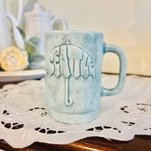 May include: A light blue ceramic mug with the word "SEATTLE" in a raised, umbrella-shaped design. The mug has a speckled finish and a curved handle. The mug is sitting on a white, lacy doily.