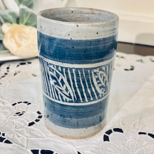 May include: A cylindrical ceramic tumbler with a speckled light gray base and dark blue horizontal bands. The center section features a hand-painted white leaf pattern against a dark blue background, creating a contrast with the speckled clay texture.