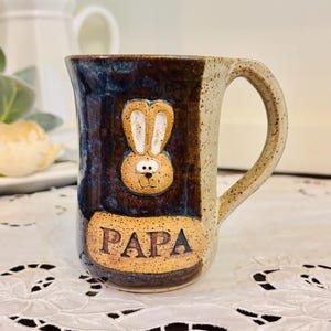 May include: A ceramic mug with a speckled cream and dark brown glaze. The front features a raised, sculpted tan rabbit face with long ears and the word PAPA printed in black capital letters on a tan rectangular patch below the rabbit.