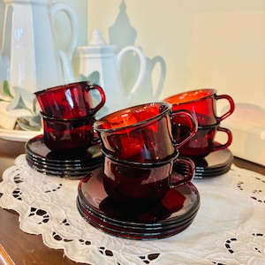 May include: A set of ruby red glass teacups and saucers are stacked on a white lace doily. The cups have a classic design with a rounded body and a small handle. The saucers are round and match the cups. White teapots are in the background.