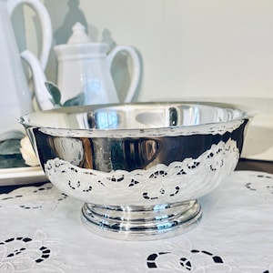 May include: A silver-colored bowl with a decorative, scalloped edge. The bowl sits on a round base and is placed on a white, embroidered doily. The background includes white teapots and a tray.