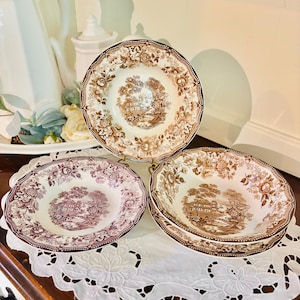 May include: A collection of vintage ceramic bowls with floral and landscape designs in shades of brown and purple. The bowls are arranged on a white lace doily, showcasing their intricate patterns and classic aesthetic.