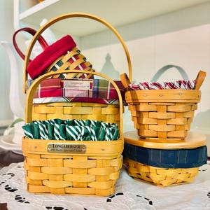 May include: A collection of Longaberger baskets in various sizes and shapes. The baskets are made of woven wood and feature fabric liners in red, green, and blue plaid patterns. Some baskets have handles, and one has a metal tag that reads "Longaberger".