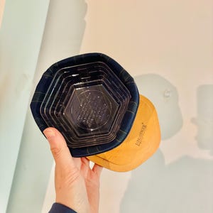 May include: A hand holding a Longaberger basket with a navy blue plaid fabric rim and a clear plastic insert. The basket has a light brown wooden base.