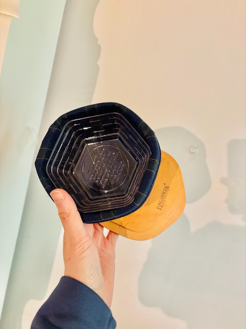 May include: A hand holding a Longaberger basket with a navy blue plaid fabric rim and a clear plastic insert. The basket has a light brown wooden base.