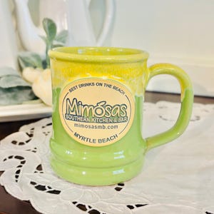 May include: A two-toned ceramic mug with a green base and yellow top. The mug features a circular sticker with the text "Mimosas Southern Kitchen & Bar Myrtle Beach". The mug has a curved handle.